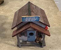 Front view of wooden bird house with brown slanted roof and 'PRO SHOP' sign. Light blue door with miniature golf bag and clubs by entrance.