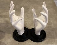 Photo shows two white hand sculptures mounted on a black base, positioned on a beige surface, highlighting the remote control holder design.