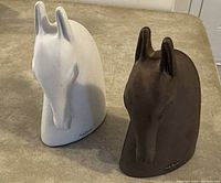 Photo shows two ceramic horse head bust sculptures, one white and one bronze, standing side by side on a beige surface. The busts have a minimalistic, smooth design with artist's signature on the base.