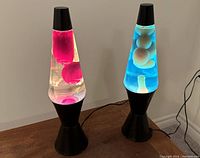 Two lava lamps side by side on a wood table, both turned on. The left lamp has red lava and clear liquid, the right lamp has blue liquid with white lava. Both have black bases and caps.