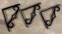 Photo of three black metal Knape & Vogt colonial style decorative shelf brackets with scroll and diamond shaped cutouts, arranged on a floor.