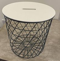 Black metal wire basket with white oval lid on top, placed on carpeted floor.