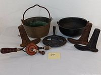 Full lot showing all items on white surface: copper cooking pot, black cast iron pan, cast iron trivet, three cobbler shoe molds, hand drill with wooden handle and orange gear wheel.