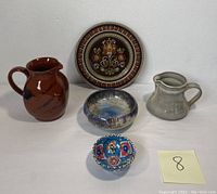 Five artisan pottery pieces including a decorative plate, brown pitcher, gray creamer, crystalline glaze bowl, and a small floral bowl on white background