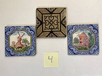 Three ceramic tiles displayed side by side on a wall, two with windmill scenes and one with an abstract pattern in black and gold.