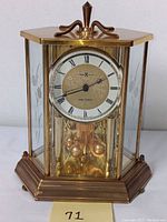 Front view of Howard Miller Mega Quartz anniversary clock showing hexagonal brass-tone case with glass panels and Roman numeral dial