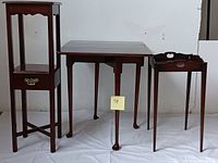 Front view of 3 pieces of mid-century wood furniture: a small stand with drawer and glass front, a drop leaf table in middle, and an end table with carved handles.