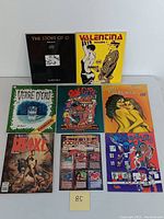 Top-down photo of eight vintage adult erotic comic books and magazines arranged in two rows, visible titles include Valentina Volume 1, The Story of O Volume 2, Verre d'Eau #28, Real Girl #1, Erotic Fantasies/Nights Children, Dangerous Pushy, and XXXenophile #10.