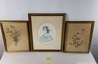 Three paintings shown side-by-side: two floral paintings on left and right, and one fashion portrait in center. All framed and under glass.