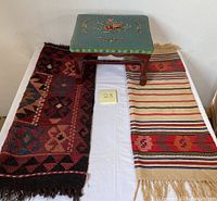 View of entire lot showing wooden stool with teal embroidered cushion and two rugs, handwoven tapestry and Afghani rug, on either side