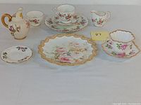 Overview of various porcelain items arranged including plates, cups, creamer and platter with floral designs and gold edging.