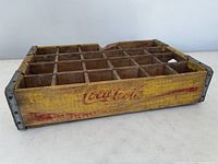 Front angled view of the yellow wooden Coca Cola crate with visible red faded Coca Cola logo and 24 compartments.