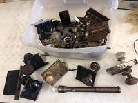 Container with assorted vintage door knobs, lock plates, hinges and a telescoping door viewer, showing a mix of brass and steel parts.