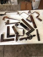 Overview of antique hand tools spread out on a white surface showing saws, braces, squares, and files in used condition.