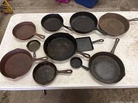 Nine cast iron pans of varying sizes arranged on a table, showing an overview of the collection and condition with some surface rust and wear.