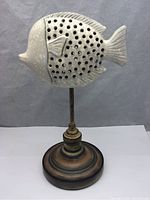 Fish-shaped decor piece mounted on a tall slender rod and round wooden base showing overall shape and perforations on body.