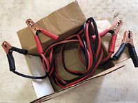 Photo showing the jumper cables coiled and placed inside a cardboard box, displaying red and black insulated clamps and cables.