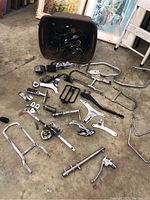 Photo of various Harley Davidson motorcycle parts spread on the floor including wiring inside a black seat bucket, chrome foot pegs, brackets, and mounting hardware.
