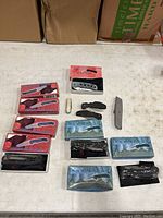 Photo of pocket knives and cutting utensils, boxed and unboxed, laying on surface with cardboard boxes in back