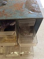 Close-up photo showing rusty and oxidized metal storage case with small transparent drawers partially open containing various tumbled stones.