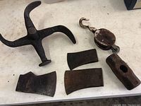 Top view of five cast iron tools on a white surface: one large four-armed star-shaped tool, one block and tackle pulley with hook, and three axe heads of varying shapes and sizes.