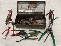 Top-down view of the small metal toolbox open showing rust and various tools inside, including pliers and wire cutters spread around the toolbox on a white surface.