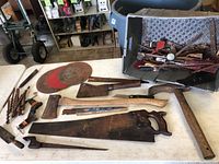 Photo showing assortment of vintage tools including hand saws, axes, circular saw blades, drill bits, and other tools on table and in box.