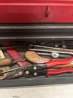 Close-up of one opened drawer showing red-handled wire cutters, pliers, small saw with wooden handle, and metal file with wooden handle inside a red metal toolbox.