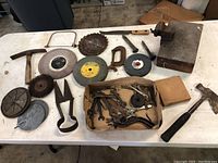 Table with a collection of various hand tools including hammer, hand saw, cutting discs, vise clamp on wooden block, metal shears, and a cardboard box with smaller tools and parts.