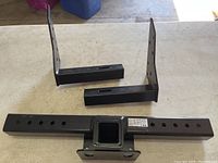 Three black powder-coated metal parts of an adjustable Reese Tow Power trailer hitch laid on a table: one rectangular receiver bar with holes and two mounting brackets.