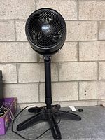 Front angled view of black Vornado pedestal fan showing the fan head, grille, and base with cords, set against a brick wall.