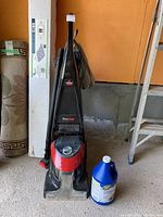 Full view of Bissell ProHeat upright rug cleaner standing upright next to a blue jug of Zep carpet shampoo concentrate on a concrete surface in a garage or storage area.