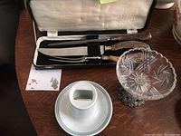 Glass pedestal dish with metal base, ceramic vintage matchbox holder, and carving set case on table.