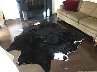 Black and white cowhide rug spread on dark wood floor near a beige couch and black chair in living room, showing white patches around edges and overall black fur.