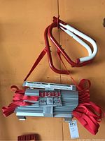 Folded aluminum ladder with red straps and instructions label visible, hanging against an orange background.