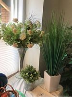 Set of three faux plant arrangements including tall glass vase with flowers, white ceramic vase with grass, and small metal bucket pot with greenery, shown near window.
