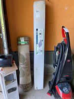 Photo of the white box packaging containing the hammock beside a rolled rug and vacuum cleaner.