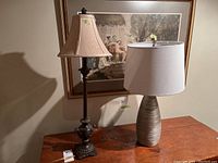 Two table lamps side by side on wooden table. One with dark ornate composite base and beige bell fabric shade, other with light textured composite base and large beige cylindrical shade.