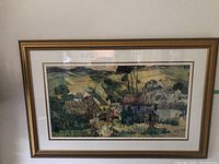 Full view of framed landscape print showing rural scene with houses and trees in green and earth tones, in gold frame with glass and matting.