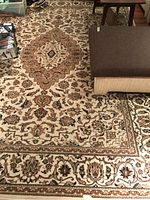 Photo showing the rug partially covered by ottomans and a small table, displaying the traditional floral pattern and central medallion design.