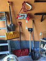 View of yellow plastic snow shovel with black handle and D-grip, red plastic snow shovel, metal shovel, and other tools hanging against a wall.
