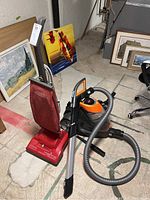 Photo showing both the Hoover Encore Supreme upright vacuum in red with cloth bag and the Bissell Multisurface Professional bagless canister vacuum in black and orange with flexible hose and attachments on basement floor near paintings.