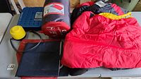 Photo showing two compact Outbound comfort sleeping bags, red and black, yellow foot pump, part of air mattress, and folded black bag on white table.