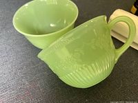 Two light green glass cups in the Fire King Jadeite Alice pattern, showing embossed floral and grape designs with handles.