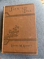 Front cover of a brown hardcover book titled 'Jack and Jill' by Louisa M. Alcott with floral line decorations.