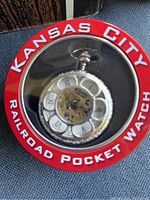 Top view of silver-tone pocket watch in original red round metal box with clear window, showing the watch face with visible gears and Roman numerals.