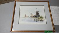 Framed watercolor painting showing three Dutch windmills by the water with reflections, framed in a wooden frame with beige mat.