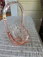 Pink depression glass basket with tall basket weave style handle and scalloped edges, sitting on a wicker surface.