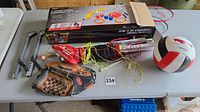 Five-game combo set box with game pieces like lawn darts and rings displayed on the box illustration, along with assembled physical items including volleyballs, baseball glove, skate guards, badminton rackets and net parts on a table.
