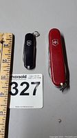 Both black and red Swiss Army pocket tools shown side by side for size comparison with measuring ruler
